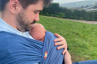 Connor looking down at his newborn daughter in a sling, with a field, trees and rolling hills in the distance.